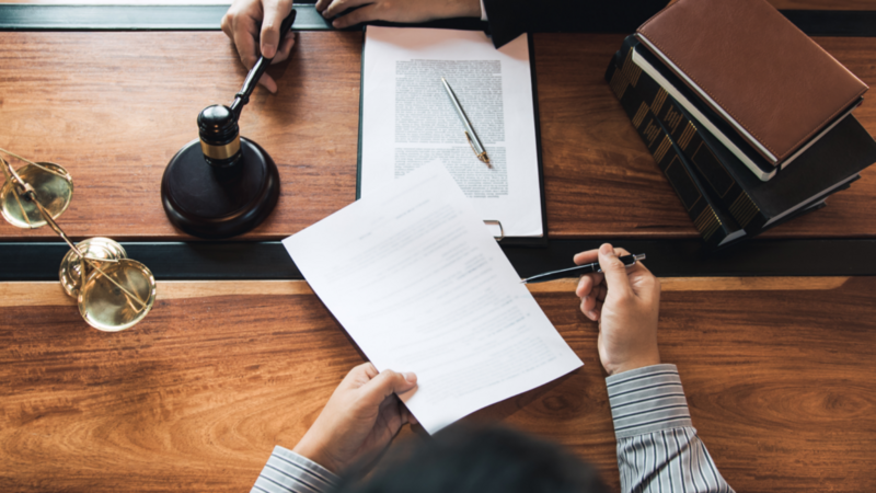 Hands of a lawyer and client reviewing legal documents on a wooden table with a gavel, scales, and law books, conveying a serious tone.