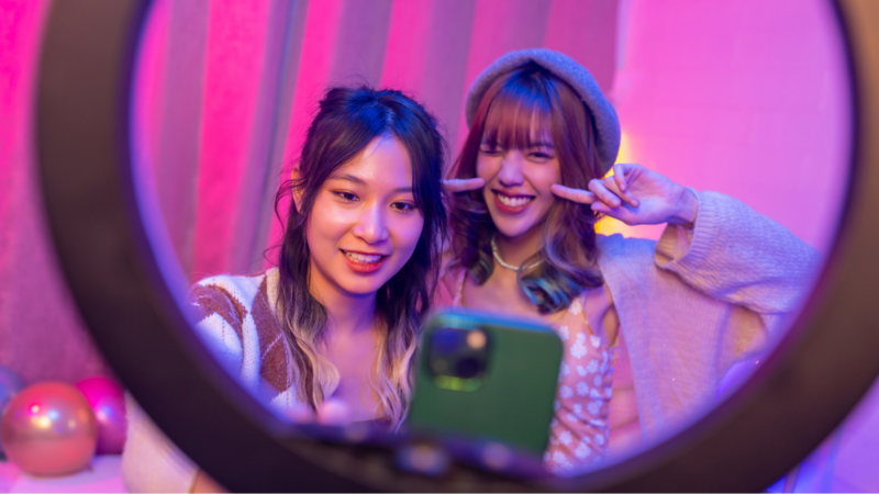 Two young women taking a selfie together in a brightly lit, colorful room, with a ring light in the foreground.