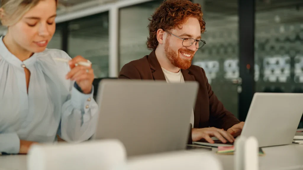 A man and woman sit together at a table, both working on laptops, immersed in their tasks.