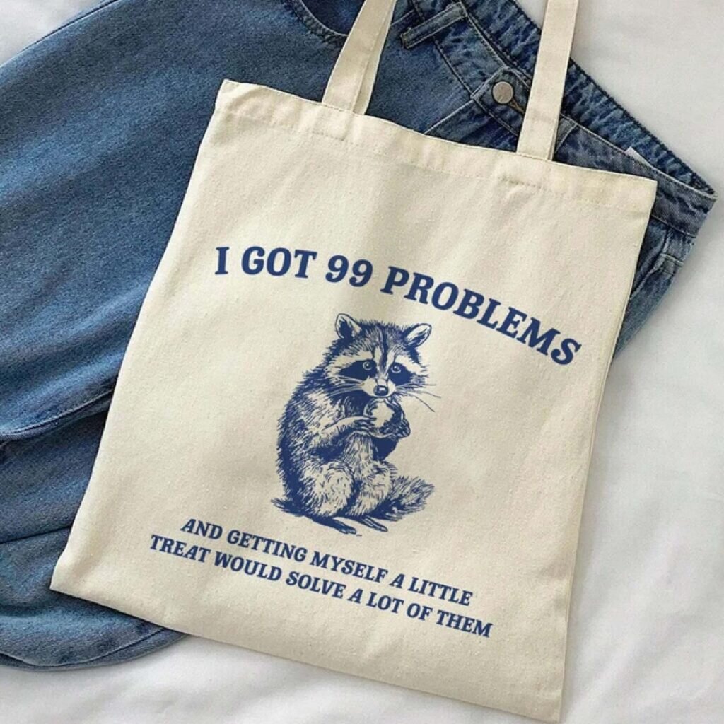A tote bag with an image of a raccoon and the caption, “I got 99 problems, and getting myself a little treat would solve a lot of them.”