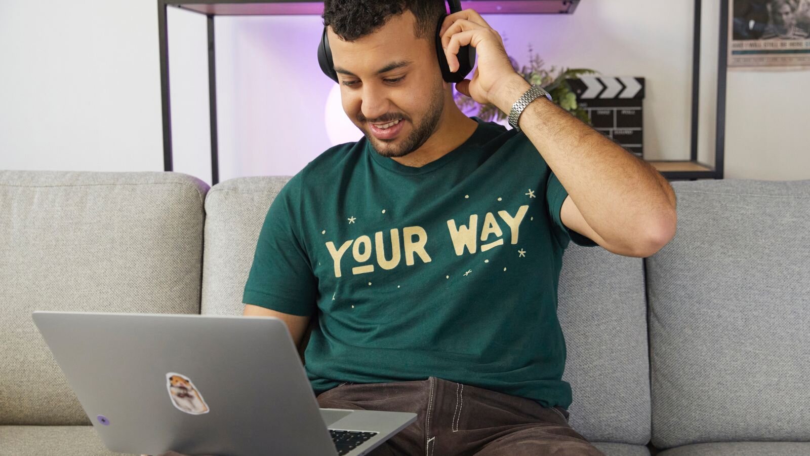 A smiling man is about to take off his headphones while looking at a laptop, learning how to sell merch on Shopify.