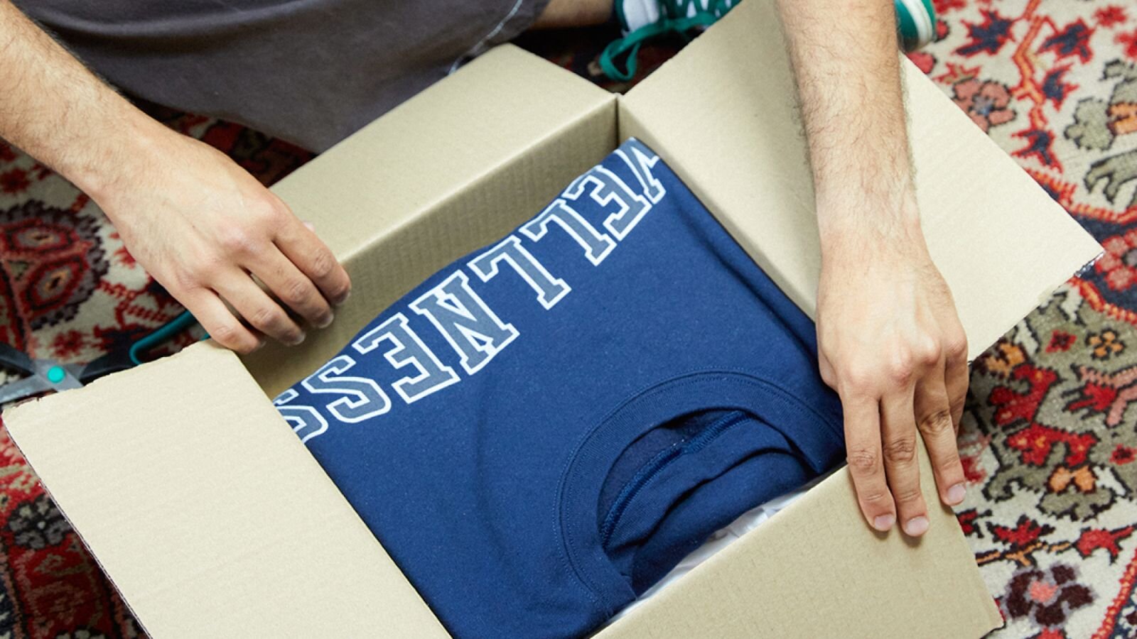A person is placing a blue t-shirt with the word "WELLNESS" printed on it inside a cardboard box.