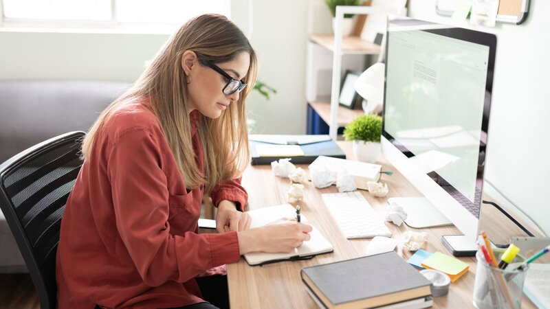 Woman at a desk writing in a notebook beside a desktop computer, surrounded by crumpled paper drafts.