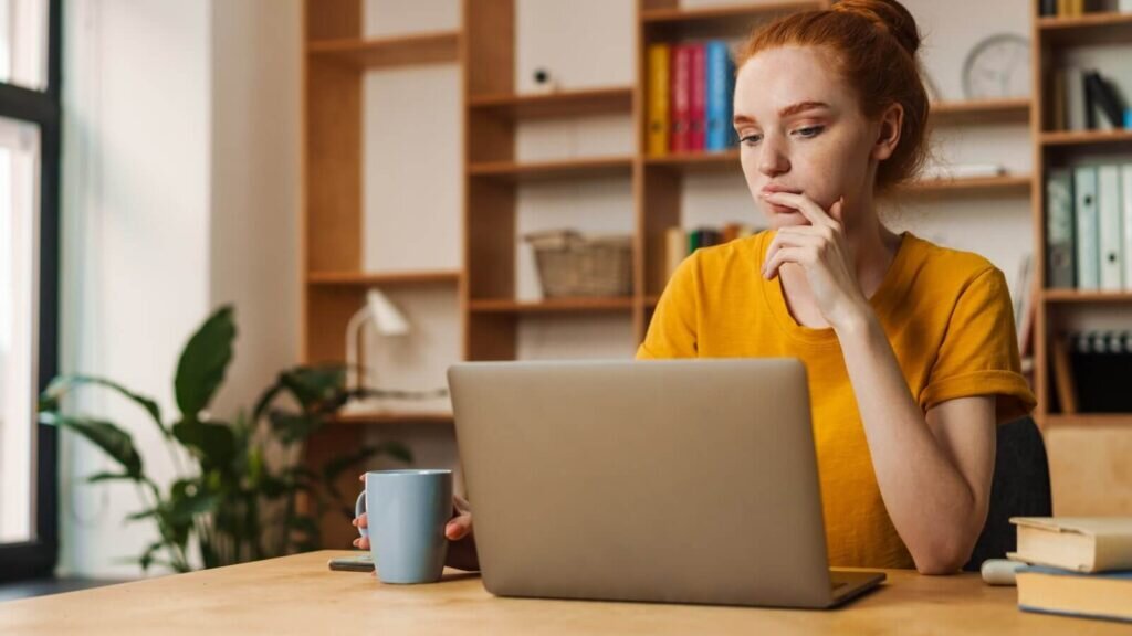 A woman sitting at a desk and looking at a laptop screen, thinking.