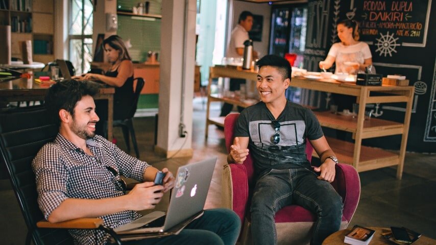 Two men laughing and sitting, one is with laptop.