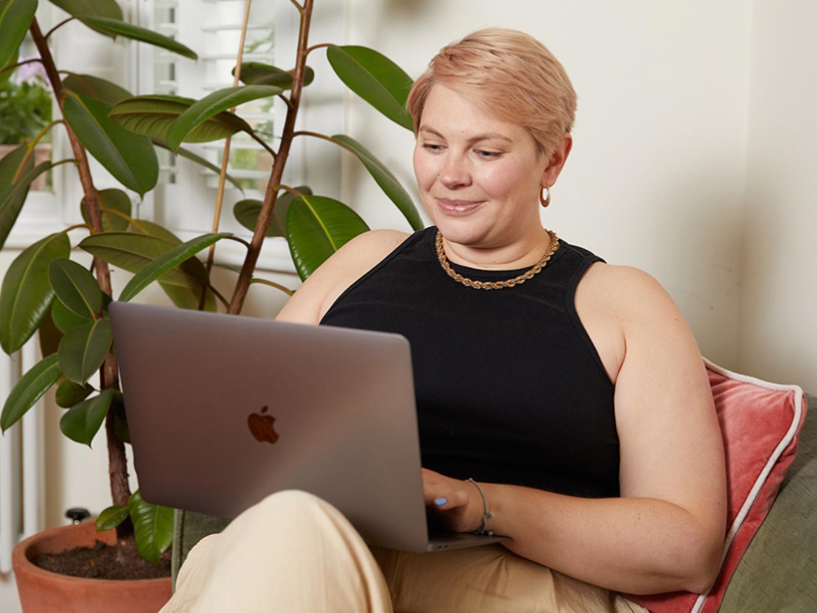 A young smiling woman with short hair sits comfortably on a couch, using a laptop.