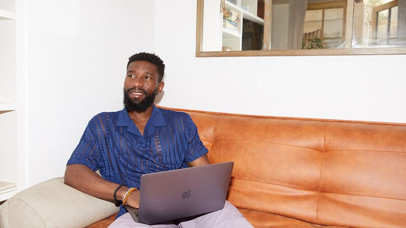 A man sitting on a leather couch with a laptop, smiling while looking to the side.
