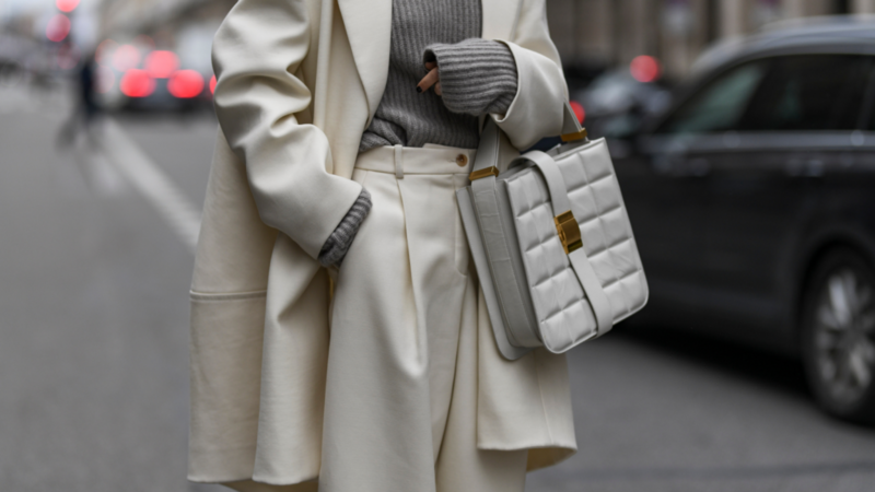 A person wearing an elegant cream coat and pants, a gray sweater, and holding a stylish quilted handbag on a city street.