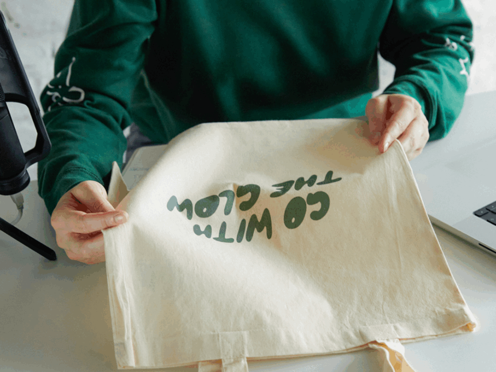 A woman in a green hoodie holding a custom “Go with the glow” tote bag.