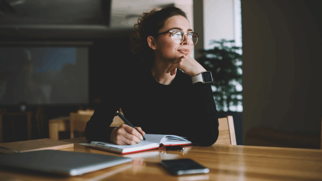 A woman wearing glasses sits at a table, writing in a notebook with a pen.