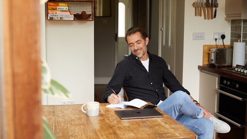 A man sitting at a table, smiling and writing down something in a notebook with a laptop beside it.
