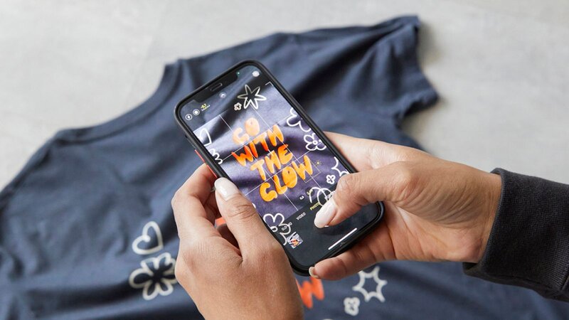 A person is taking a picture of a blue t-shirt, with the text "GO WITH THE GLOW", using a smartphone.