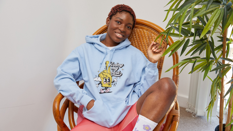 A smiling woman with short hair sitting in a wicker chair, wearing a light blue hoodie with a playful cartoon design and pink shorts.