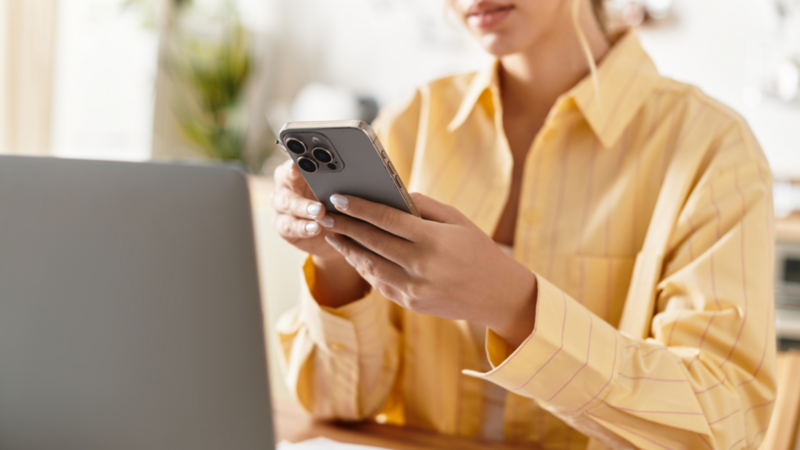 A woman in a yellow blouse is sitting at a desk and holding a smartphone in her hands in front of a laptop.