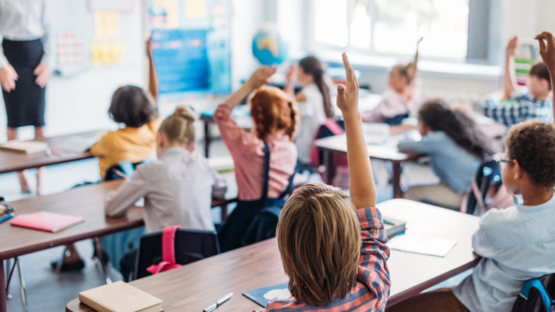 A classroom with young students sitting at desks, raising their hands eagerly.