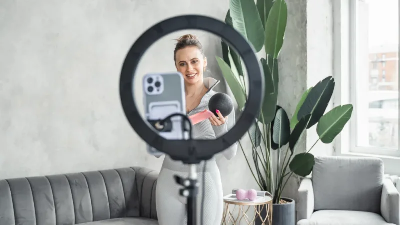 A woman in a cozy living room records a fitness vlog. Holding a pink water bottle and a black exercise ball.