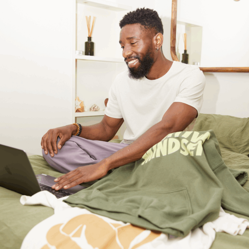 A smiling man sitting in bed with a laptop on his lap, and a “Positive mindset” hoodie next to it.