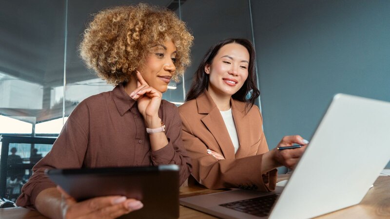 Two women in an office are actively discussing, with one woman holding a tablet and the other pointing at a laptop screen.