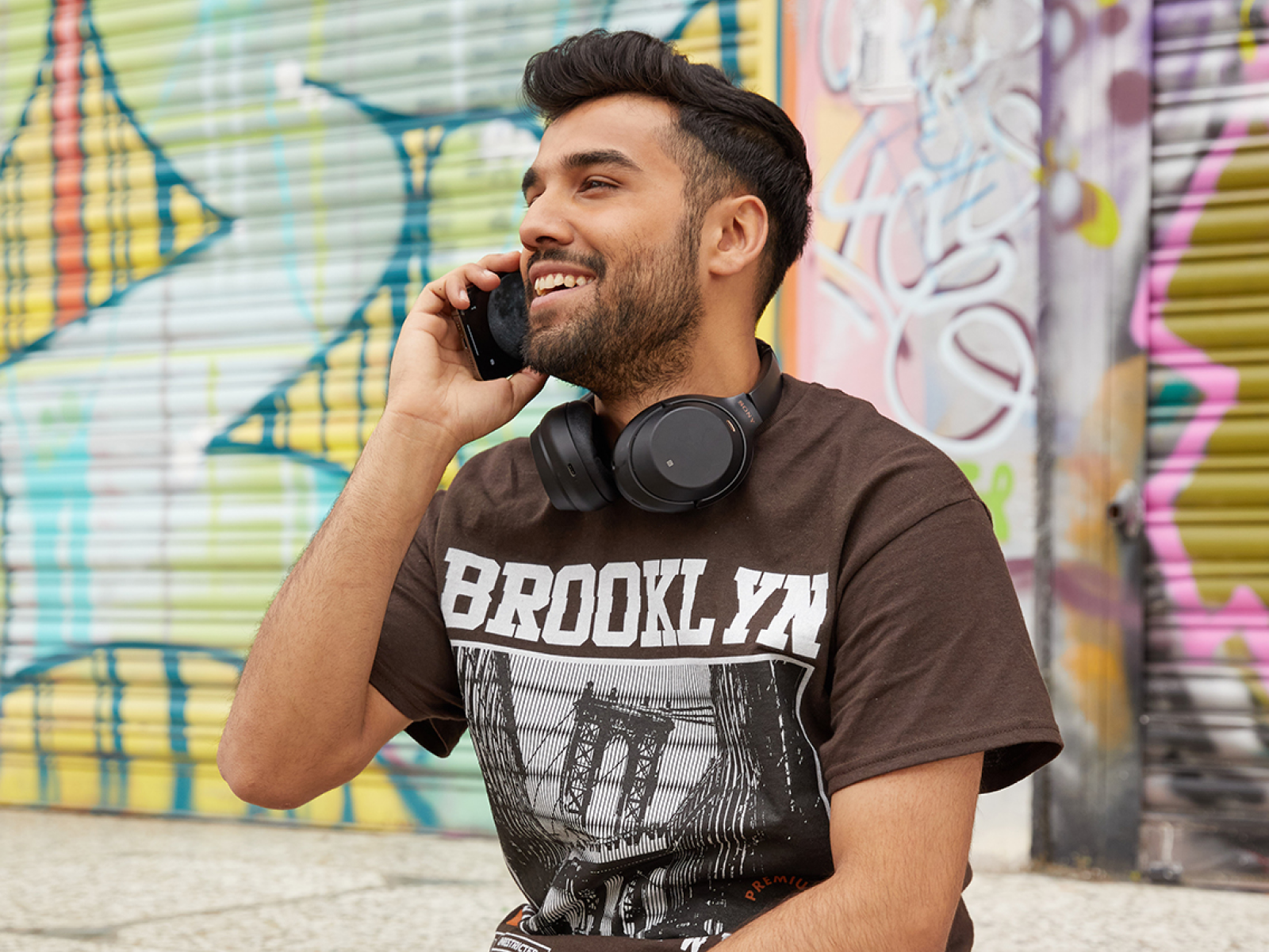 A man talking on a phone, wearing a brown Bella Canvas 3001 “Brooklyn” t-shirt.