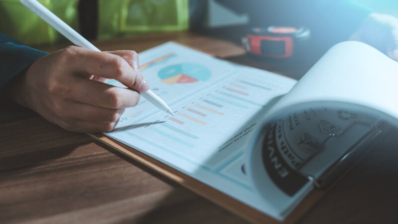Close-up of a hand using a stylus to review a printed bar and pie chart report on a clipboard.
