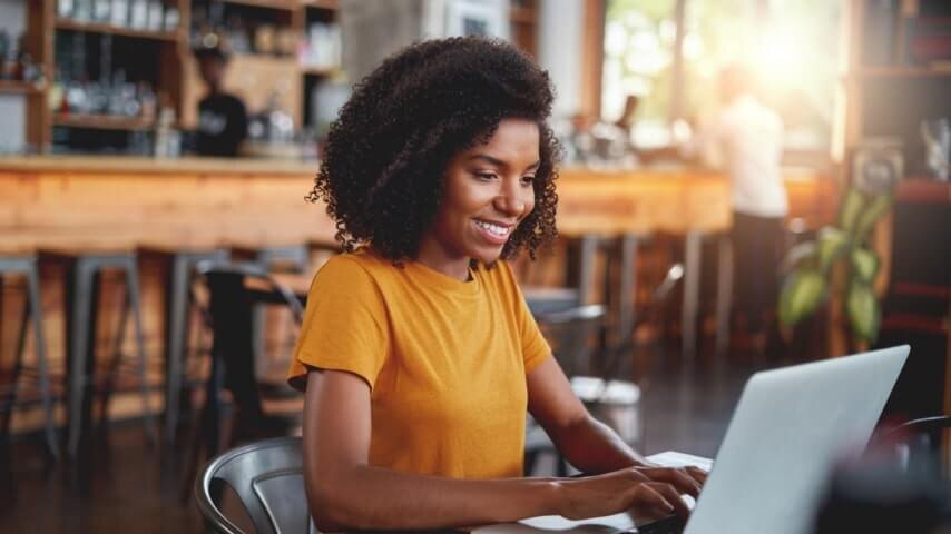 A woman in a yellow t-shirt looking at a laptop screen and smiling while typing something.