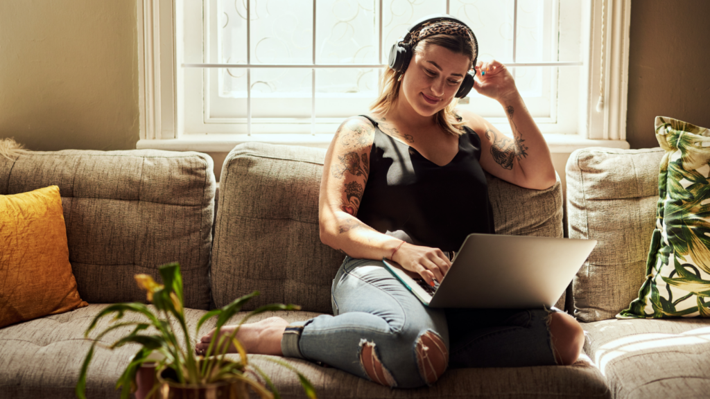 A smiling woman in headphones is sitting with a laptop on her lap, using Printify’s Product Creator.