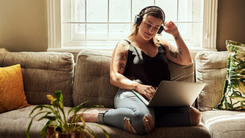 A smiling woman in headphones is sitting with a laptop on her lap, using Printify’s Product Creator.