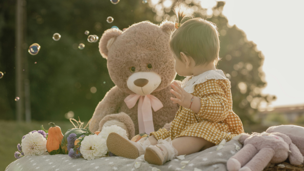 A baby girl seated on a blanket, with a teddy bear resting next to her.
