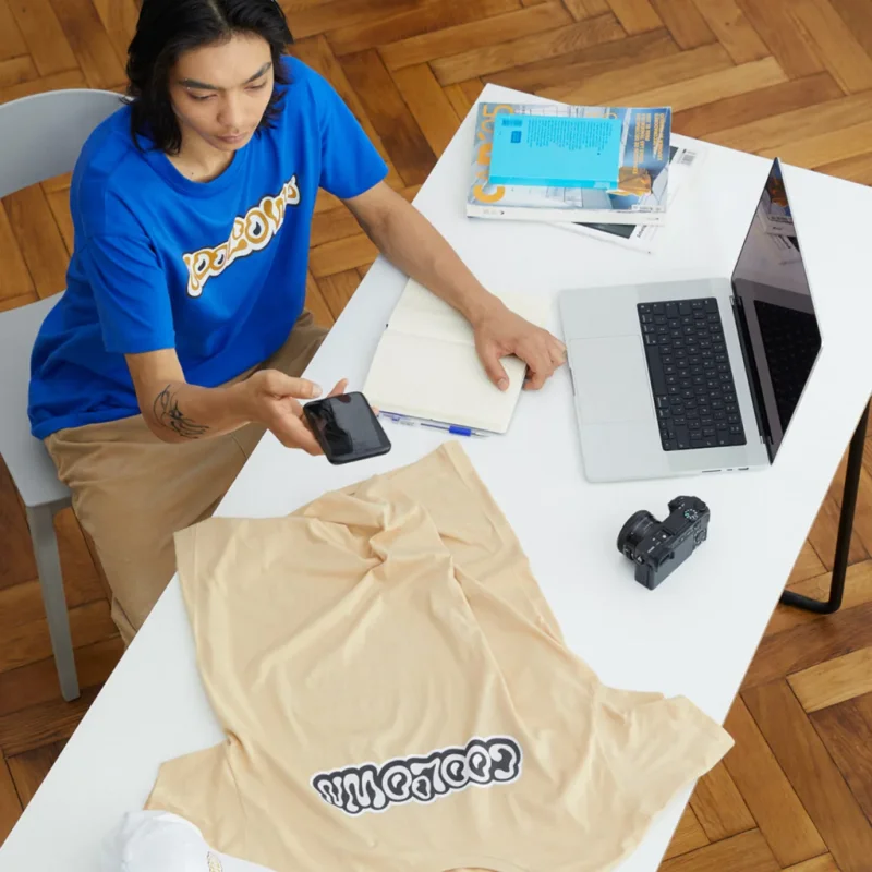 A man in a blue t-shirt sits at a white desk. He’s taking a picture of a beige t-shirt, surrounded by a notebook, camera, and open laptop.