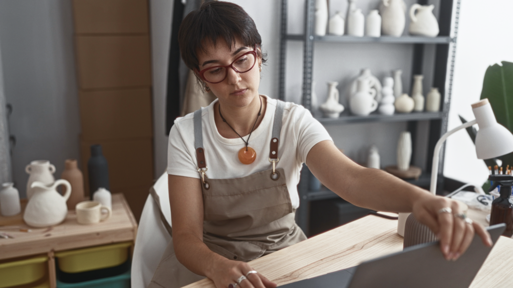 A woman with short hair, in glasses, wearing a work apron, is sitting in front of a laptop, touching it’s screen. Shelves with pottery behind her.