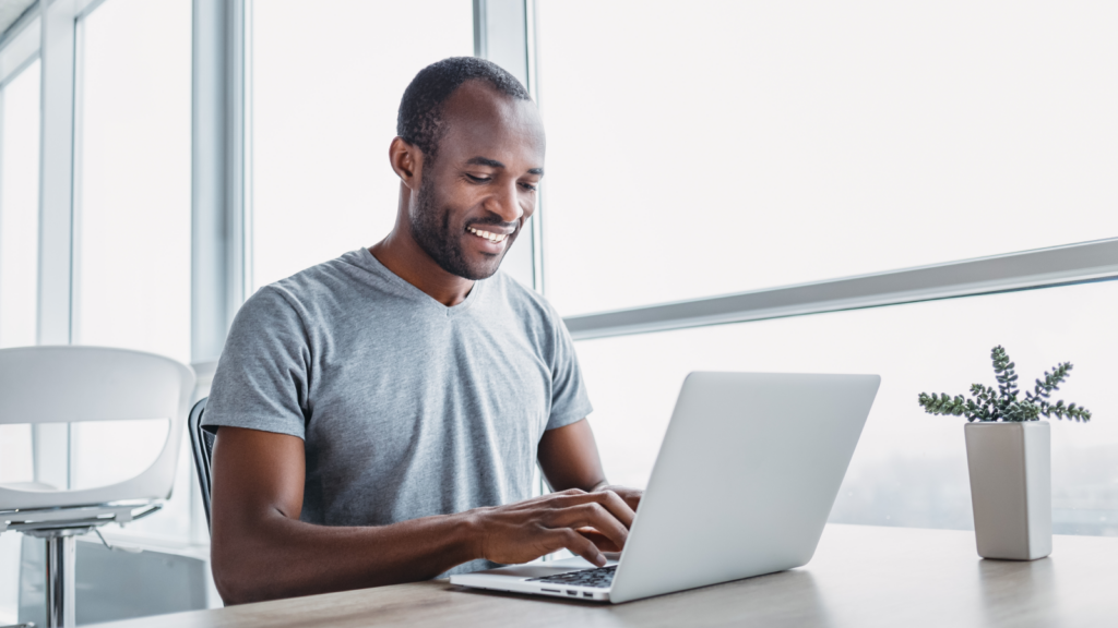 A cheerful man at a table uses his laptop, exuding a sense of happiness and engagement in his work.