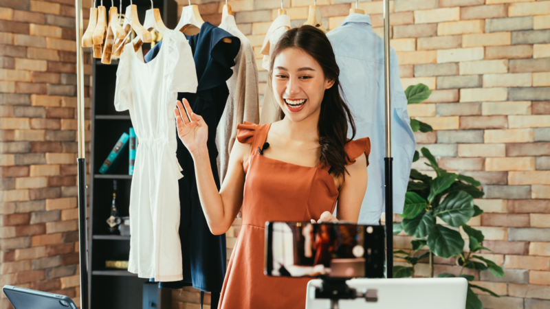 A young woman in an orange dress is smiling and waving while being recorded with a smartphone, surrounded by clothing on display.