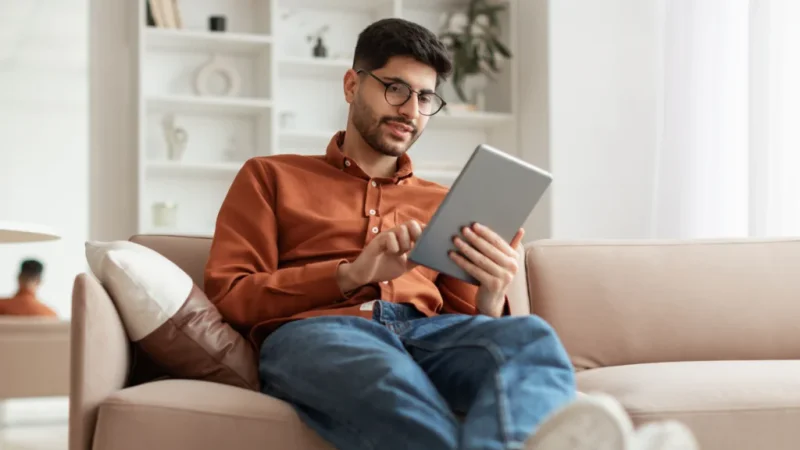 A man with glasses, wearing a rust-colored shirt and jeans, sits relaxed on a beige sofa using a tablet.