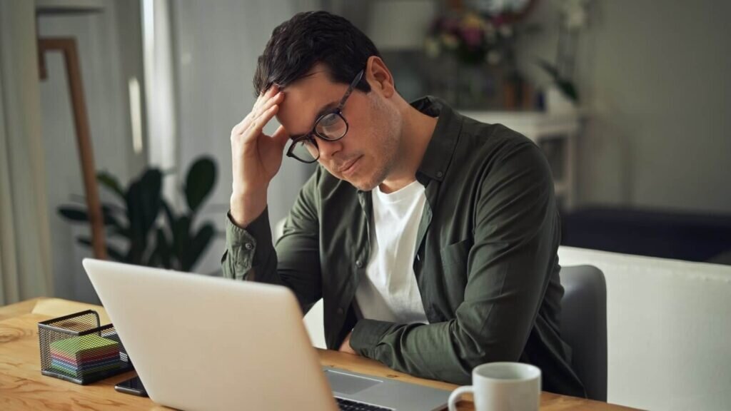 A man in a dark green shirt nodding his head to the side and thinking while looking at a laptop screen.