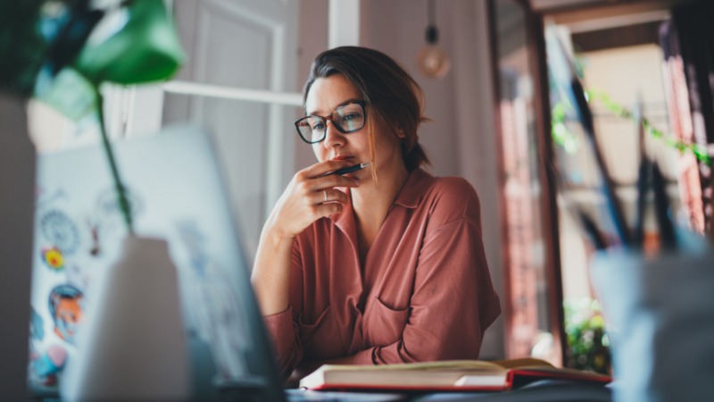 A woman in glasses and a pink shirt in a modern office looks at her laptop, learning if you can sell AI-generated art.