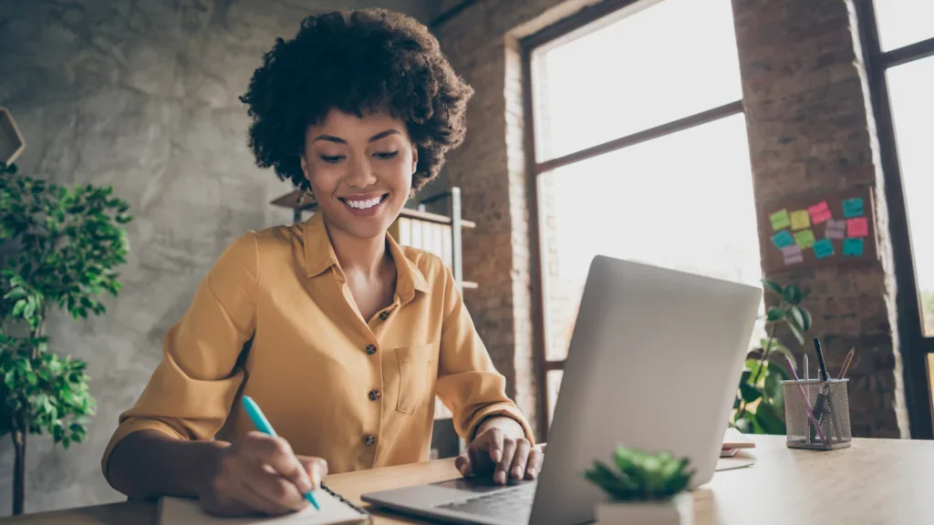 A smiling woman sits at a desk, focused on her laptop while working in a bright, inviting environment.