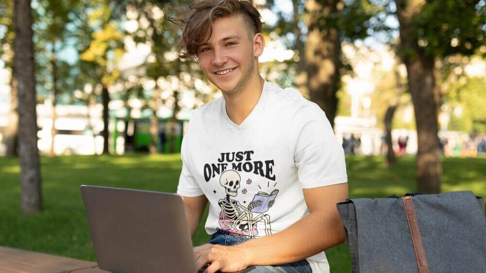 A young man sitting with a laptop on his lap, wearing a white Gildan 64V00 t-shirt, with an illustrated skeleton print.