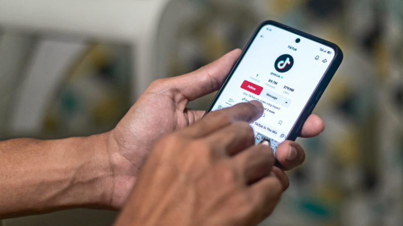 A person holds a smartphone displaying a TikTok profile with the app logo visible.
