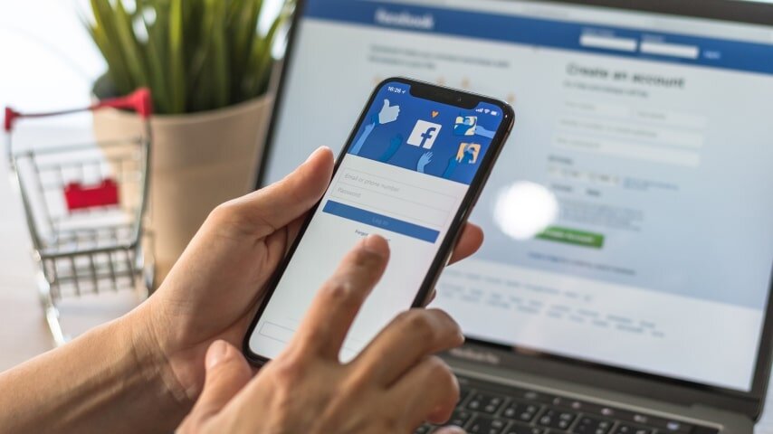 A man holding a mobile phone in front of a laptop with Facebook open on both.