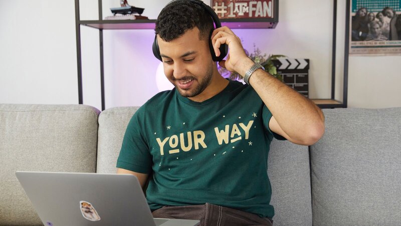 A smiling man in headphones, wearing a green “Your way” t-shirt, is sitting with a laptop, learning how to promote a t-shirt business.