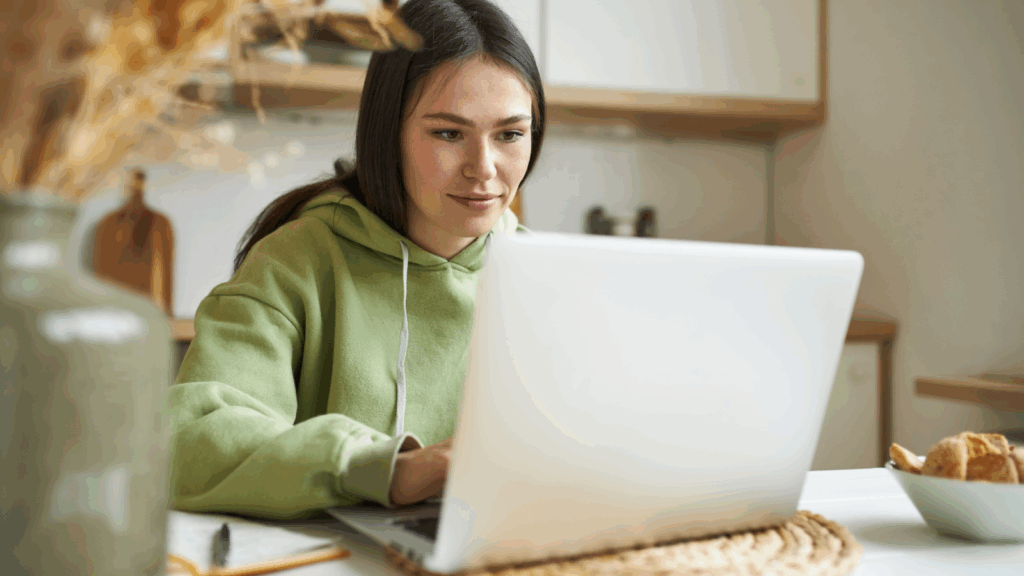 A woman in a green sweatshirt is focused on using a laptop at a desk.