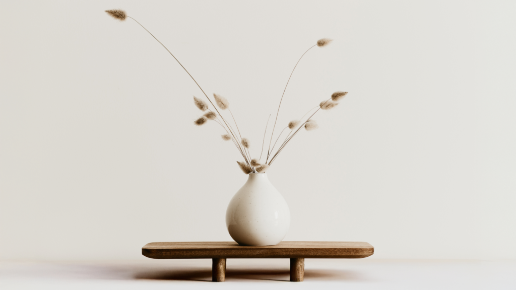 Minimalist scene with a white vase holding wispy grasses on a small wooden table.