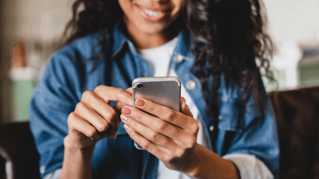 A smiling woman is using a mobile phone that she’s holding in her hands.