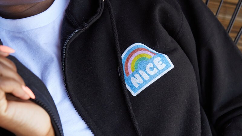 A person wearing a black zip-up hoodie with a "NICE" patch featuring a rainbow design.