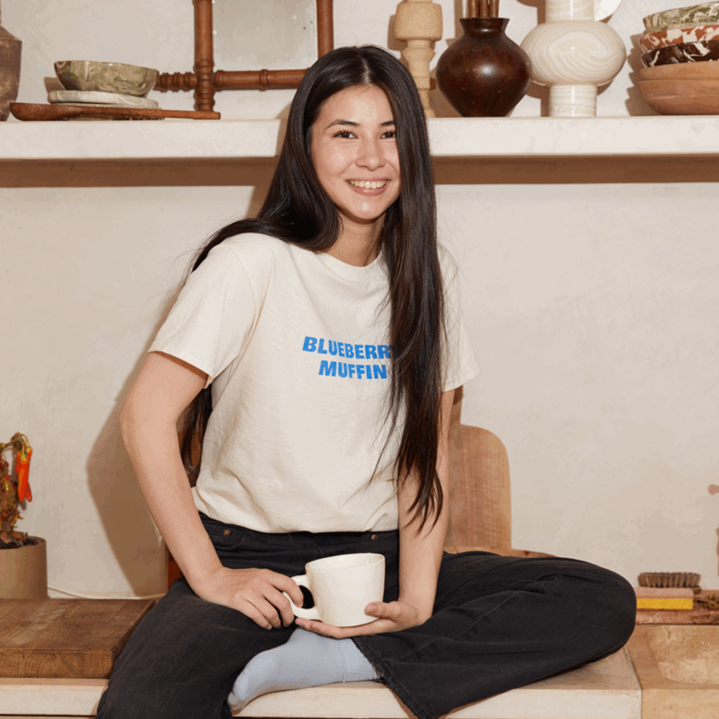 A smiling young woman wearing a beige cotton t-shirt with a blue “Blueberry muffin” text print on the front.
