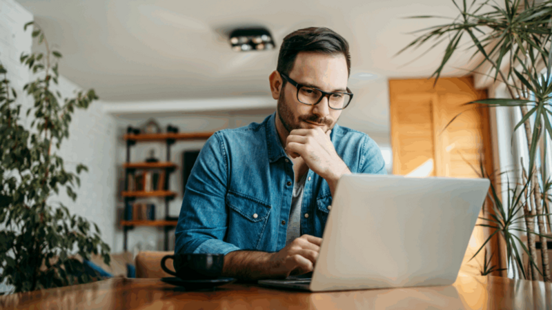 A man in glasses is sitting at a desk and using a laptop.