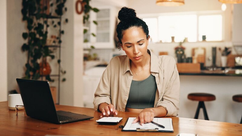 A woman with black hair, wearing a beige shirt, is sitting at a desk with a laptop, working with a calculator and making notes.