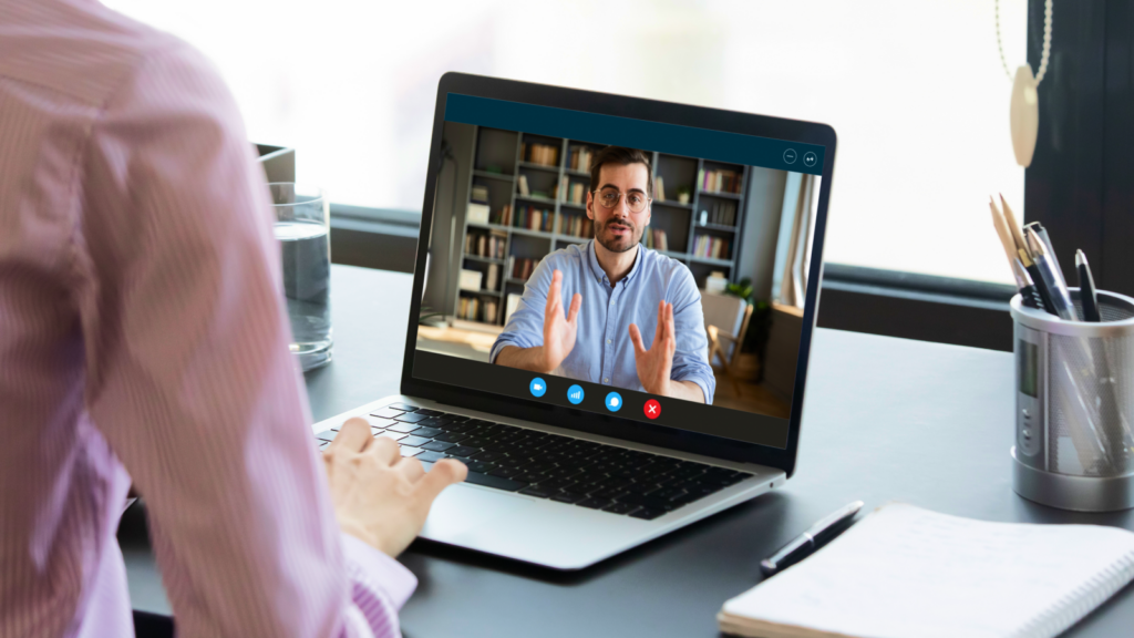 A person in a striped shirt sits at a desk viewing a video call on a laptop. The screen shows a man gesturing while speaking.