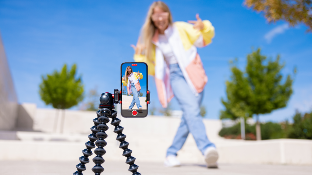 A woman stands in front of a tripod, holding a phone, preparing to capture a photo or video for social media.