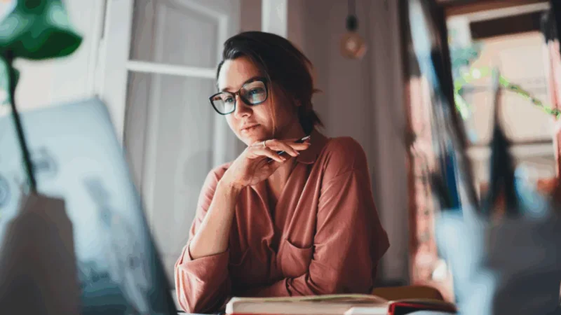 A woman wearing glasses is seated at a desk, working on a laptop.